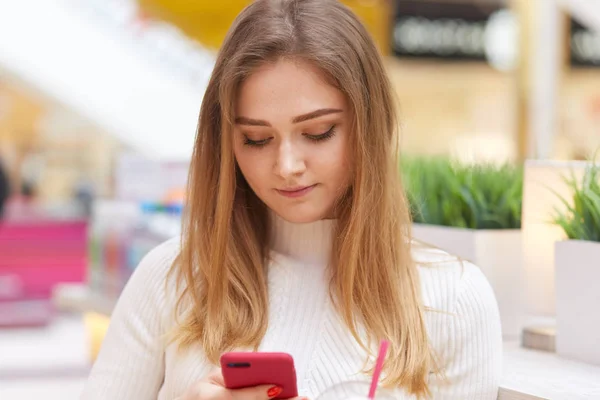 Teenage girl with long hair, focused in screen of cell phone, reads news in internet, surfes social networks, reads post under photo, poses against blurred background indoor, has spare time. — Stock Photo, Image