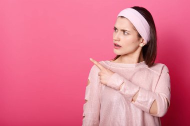 Kendine güvenen Avrupa kadın gül gömlek ve saç bandı, bir kenara Puan giymiş ve seriuosly kopya alanı olarak görünüyor. Genç Bayan şaşırtıcı bir şey görür. Reklam kavramı. Pembe arka plan izole.