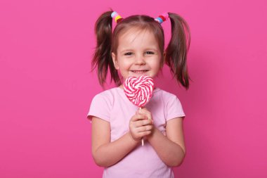 Güzel kadın çocuk tutan büyük çizgili lolipop şekli kalp, mutlu ve heyecanlı görünüyor, gül t gömlek giyer, pembe bir arka plan üzerinde izole gülümseyen duruyor. Küçük kız tatlı şeker sever.