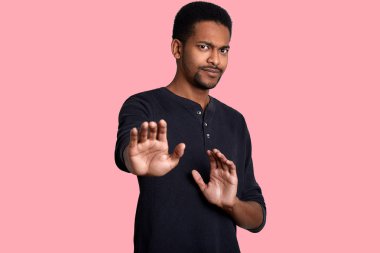 Portrait of African American male against pink wall. Guy has unpleasent conversation, strenches hands and shows to his interlocutor stop gesture, looks at the camera with worried expression. clipart