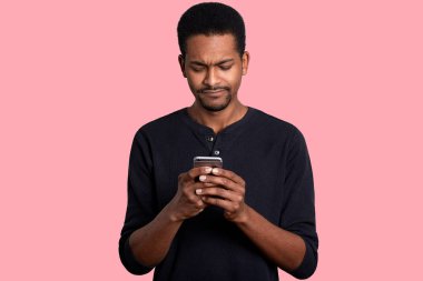 Attractive black man holds mobile phone and looks worried. Black guy reads message with unpleasent news from friend and does not know how to help. Male wears casual jumper, stands against pink wall. clipart