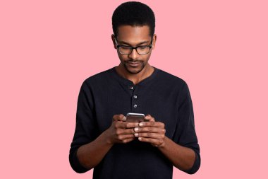 Kendine güvenen genç Afrika adam akıllı telefon görünüyor ve metni gönderir kablosuz internet'e bağlı haberci, pembe bir arka plan karşı duruyor. Koyu tenli adam siyah gömlek ve gözlük pozlar studio giyer..