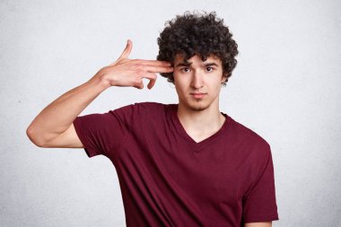 Genç yakışıklı adam rahat kıyafet giyer, kendisini öldürmesi gibi yapıyor, beyaz arka plan üzerinde izole el ile tapınağında vuruyor. Kıvırcık saçlı genç mutsuz ve üzgün görünüyor, intihar jest gösterir.