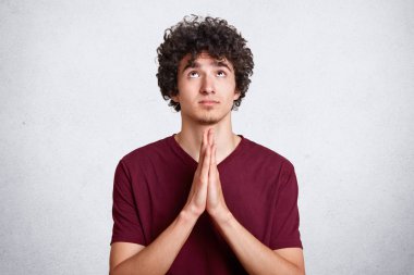 Kıvırcık siyah saçlı genç yakışıklı adamla birlikte elleriyle dua ediyor. Genç yüz, umut ile rahat bordo t gömlek elbiseler gökyüzü, arar sorar AF beyaz duvara. Din kavramı.