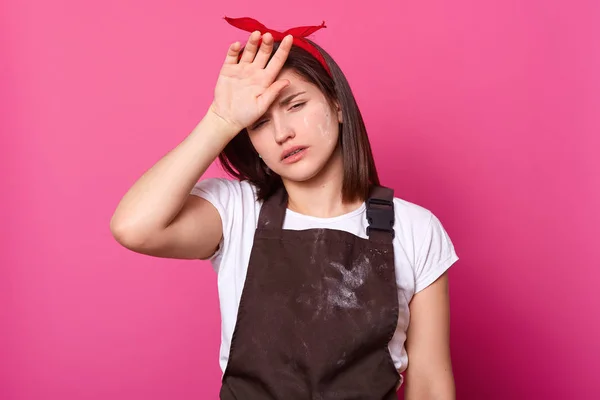 Retrato de niña en delantal marrón, camiseta blanca, banda de pelo rojo, con expresión cansada sobre fondo rosa aislado. Modelo posa en estudio fotográfico con los ojos medio abiertos, mantiene la mano en la frente . — Foto de Stock