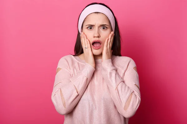 Close up photo of shocked amazing brunette with opened mouth, keeps hands on cheeks, has astonished facial expression, sees something terrible, wears rose shirt, hair band isolated on pink background.