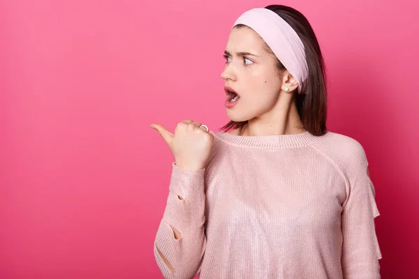 Närbild porträtt av attraktiva brunett flicka visar något åt sidan. Förvånad över kvinnan pekar vänster tumme finger och ser rädd. Kvinna med öppnade munnen står förvirrad över rosa bakgrund. — Stockfoto