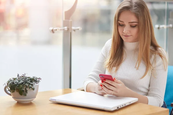 Successful long haired blonde business girl works online. Tired women wants to have rest, closses her laptop and read mesages in social network using smart phone. Young lady sits at table in cafe. — Stock Photo, Image