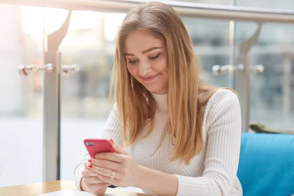 Smiling young blonde woman with long straight hair, in white shirt massages in her cellphone while sitting in cafe. Attractive girl recives sms from her boyfriend, has pleasent news. People concept. — Stock Photo, Image