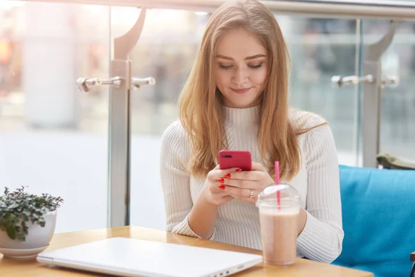 Elegáns fiatal fehér pulóver szőke nő ül a kék kanapé, kávézó, rózsaszín mobil telefon és surfes interneten vagy típusú üzenetet barátja tartja. Vonzó nő italok koktél és ellenőrizze e-mailben. — Stock Fotó