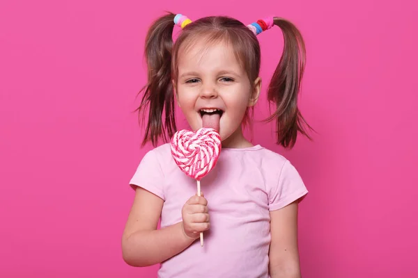 Niña feliz con piruleta rayada en forma de corazón en la mano. Charming niño lleva casual rosa camiseta, con dos divertidas colas de caballo. Lindo niño lame deliciosos dulces aislados sobre fondo rosa . — Foto de Stock