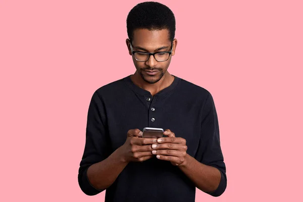 Confident young African man looks at smart phone and sends text messege, connected to wireless internet, stands against pink background. Dark skinned guy wears black shirt and glasses poses in studio. — Stock Photo, Image