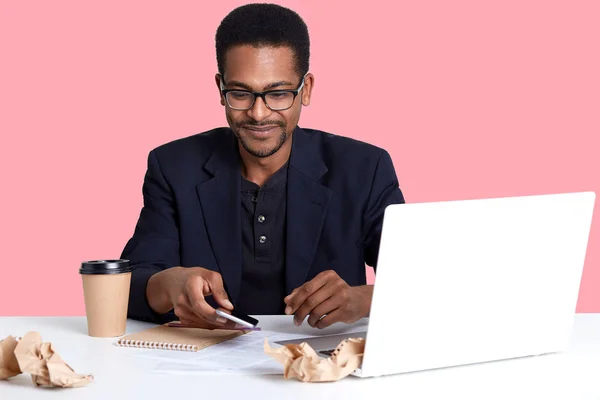 Handsome African American male sits at desk with lap top and crumpled shirts of paper, works online. Dark skinned guy takes smart phone and looks down on it, drinks coffee or tea. Technology concept.