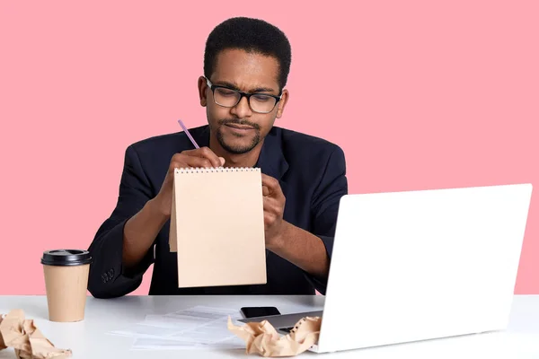 Doordachte zwarte zakenman probeert te schrijven gedicht in notitieblok voor haar vriendin terwijl u werkt aan laptop. HANDSOM African American man draagt blak shirt en jas zit tegen roze muur in studio. — Stockfoto