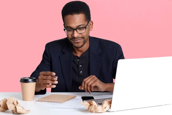 Portret van donkere gevilde man in zwarte jas en bril werkt met laptop, schrijft notities in een notitieblok en drinken koffie. Knappe zwarte man aan het bureau zit, ziet er attent. Online werk concept. — Stockfoto
