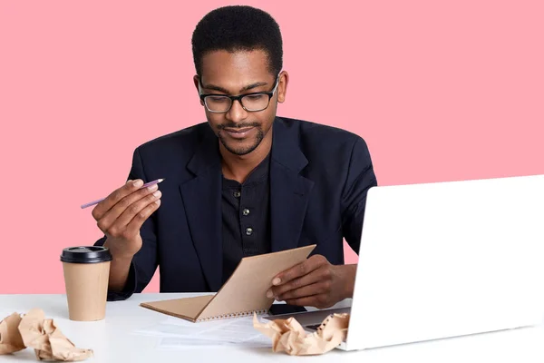 Knappe Afro-Amerikaans zakenman in jas en bril maakt gebruik van laptop. Zwarte man pen en notitieblok in handen houdt, wil opschrijven nieuw idee geïsoleerd op roze achtergrond. Bedrijfsconcept. — Stockfoto