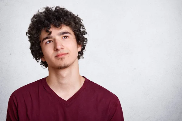 Portrait de jeune homme pensant aux cheveux bouclés foncés, se tient avec une expression faciale réfléchie, habille chemise marron. Rêves d'étudiants isolés sur un mur de béton blanc. Concept jeunesse et adolescence . — Photo