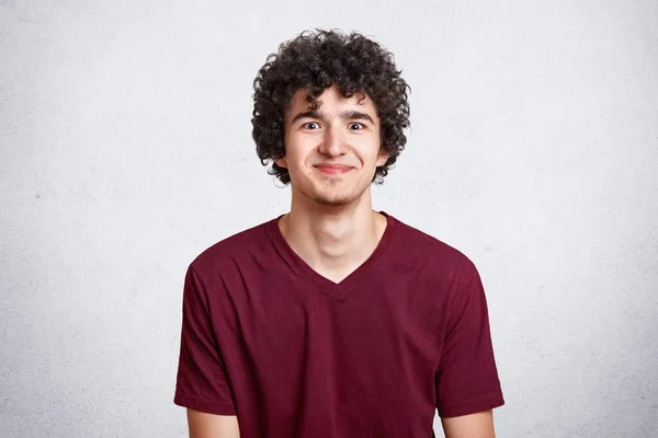 Waist up shot of young attractive guy with curly hair, dresses casual maroon t hirt, free space for your advertisment, models over white concrete wall. People, youth, clothing, advertisement concept. — Stock Photo, Image