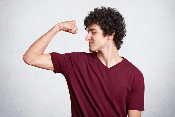 Positieve donker harige tiener, terloops, gekleed toont biceps met tevreden gezichtsuitdrukkingen, toont aan hoe cool hij is. Student met krullend haar kijkt naar zijn muscules. Sport en gezond concept. — Stockfoto