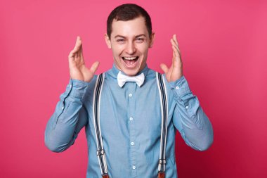 Memnun emin şık yakışıklı stüdyo portre kadar kapatın. Şanslı adam eller hapily yukarı yükseltir. Genç erkek WINS lotery, bir şey sevinçle bağırır. Gül arka plan üzerinde izole modeli pozlar.