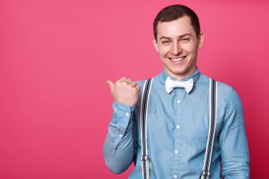 Pembe arka plan karşı ayakta önerir kullanmak kopya alanı için logo, mutlu gömlek, pantolon askısı ve papyon, yakışıklı delikanlı uzağa işaret başparmak parmak ve gülümseyen komik ifade sahipken.