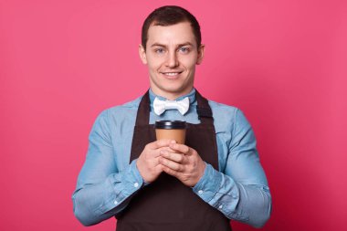 Çekici erkek barista mavi tişört, beyaz papyon giyiyor ve samimi bir gülümseme, Garson pembe arka plan üzerinde izole kahverengi önlük, tutar kağıt bardak kahve, vardır. Reklam için yer kopyalayın. İçki kavramı.
