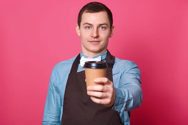 Junge hübsche männliche Barista schlägt Ihnen eine Tasse Kaffee von ihm gemacht, gekleidet elegant blaues Hemd, weiße Fliege und braune Schürze, isoliert über rosa Hintergrund. Kopierraum für Ihr Logo. Getränkekonzept. — Stockfoto