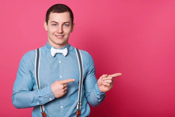 Portret van de jonge mens in blauw shirt, jarretels en wit strikje, toont beide wijsvinger opzij, geïsoleerd op roze achtergrond in de studio. Knappe Glimlachende man met aangename gezichtsuitdrukkingen. — Stockfoto