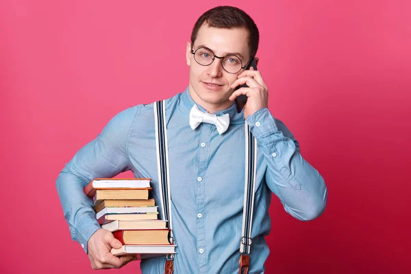 Joven adolescente masculino con expresiones faciales agradables, mantiene gran pila de libros, habla por teléfono, se prepara para las clases en la universidad, le gusta descubrir algo nuevo, aislado en el fondo de rosa . — Foto de Stock