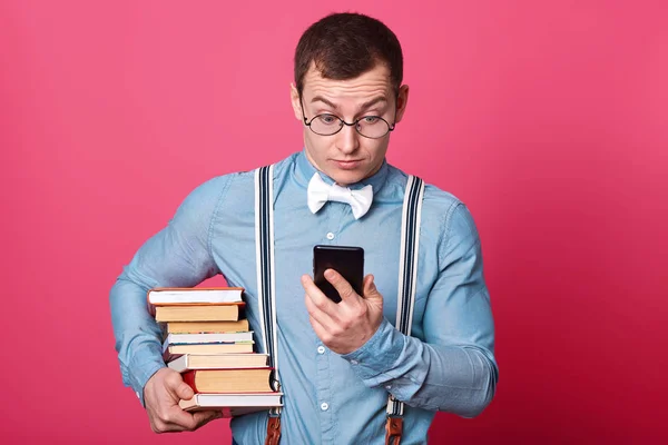 Sorprendido estudiante con pila de libros sostiene su teléfono móvil, mira la pantalla con sorpresa. Joven hombre lleva camisa, tirantes, pajarita y gafas posa en estudio aislado sobre fondo rosa . — Foto de Stock
