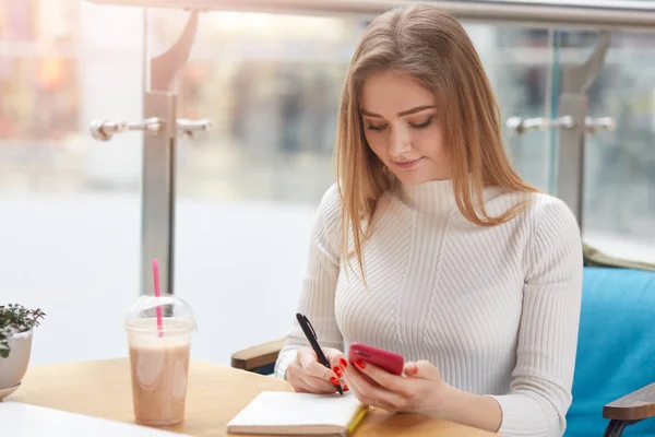 Beautiful Caucasian blonde girl sits at table, makes some notes in cafeteria, prepasres for exam. Charming student in white sweater visits coffee shop, holds smart phone in hand. Students concept. — Stock Photo, Image