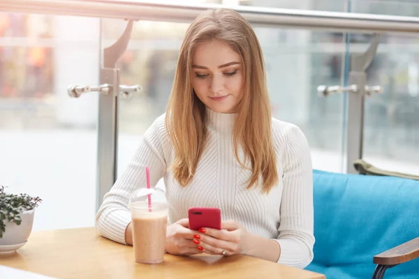 Sevimli sarışın kadının fotoğrafını akıllı telefon kullanan, kafe, surfes sosyal ağlar kanepede oturuyor, arkadaşına metin iletisi gönderir. Sevimli kız pozlar Restoran pencere üzerinde izole. Teknoloji kavramı. — Stok fotoğraf