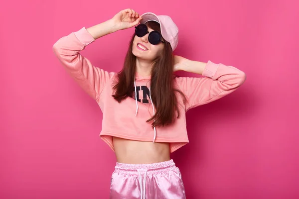 Portret van gelukkig lachend Kaukasische brunette meisje in stijlvolle korte hoodie, trainingsbroek en roos cap, draagt zwarte zonnebril, houdt hads op hoed. Vormt tegen roze achtergrond. Tieners concept. — Stockfoto
