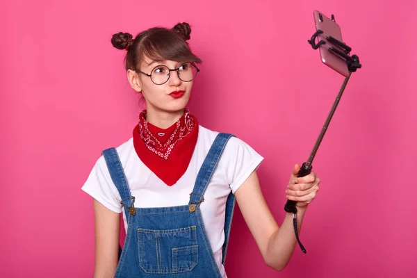 Hermosa morena femenina en camiseta blanca casual, overoles, bandana roja en el cuello y gafas redondas. Adorable adolescente mantiene los labios redondeados, hace foto con palo selfie, aislado sobre fondo de rosa . — Foto de Stock