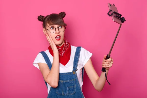 Primer plano retrato de mujer atractiva haciendo selfie, usando palo selfie, posa, toma fotos para su red social, hace mirada sorprendida, se para con la boca abierta, mantiene la mano en la mejilla. Concepto adolescente . — Foto de Stock