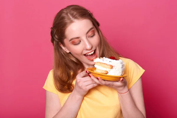 Foto de una joven hembra en una camiseta amarilla casual, lista para comer un enorme pedazo de pastel, mira la pastelería sabrosa, aislada sobre fondo de rosa, retrato de estudio. Culinario, personas y concepto de estilo de vida . —  Fotos de Stock