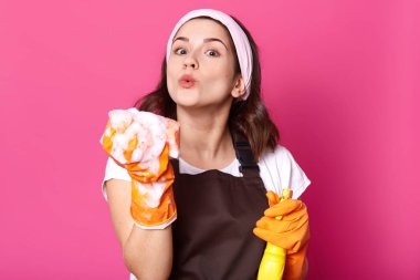 Görüntü hoş yüz ifadesi olan genç t gömlek ve önlük giyer, sprey tutan ev işi yaparken mutlu görünüyor ve köpük sünger darbeler. Hijyen, temizlik ve temizlik kavramı.