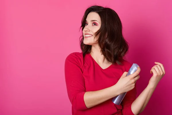 Hermosa chica de colocación fija el uso de laca para el cabello, se prepara para salir con un amigo varón, hace nuevo peinado. Fresado morena posa aislado sobre fondo rosa, mira a un lado, sostiene la botella de moussein manos . —  Fotos de Stock