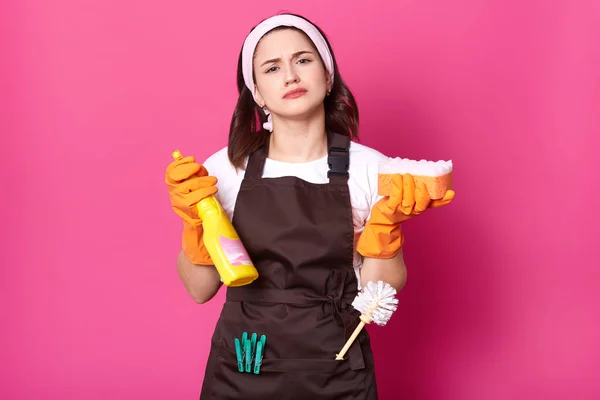 Potret wanita muda Kaukasia dengan kemeja kasual putih, celemek coklat dan pita rambut, menyimpan spons dan deterjen pembersih, terisolasi di atas latar belakang studio merah muda, siap untuk melakukan pekerjaan rumah tangga — Stok Foto
