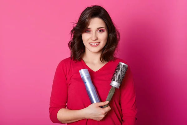 Imagen de peluquero femenino sostiene peine y hairspay, lleva suéter rojo, listo para hacer el peinado de su cliente, modelos sobre fondo rosa. Copia el espacio para el texto promocional o la publicidad. Concepto de belleza . — Foto de Stock