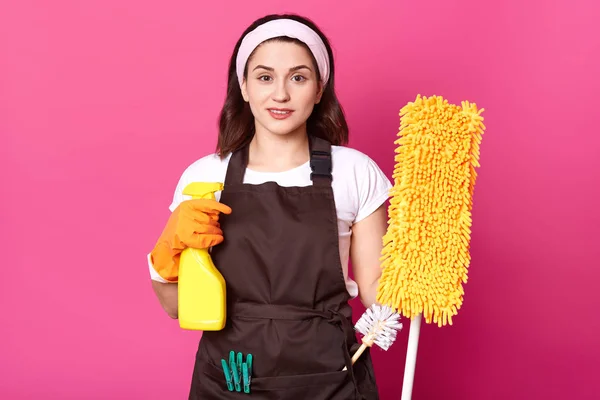 Young woman holding yellow mop and detegent spray, preparing for cleanini her house, has pleasent facial e[pression, dressed white t shirt, brown apron, isolated on rosebackground. Hygiene concept. — Stock Photo, Image