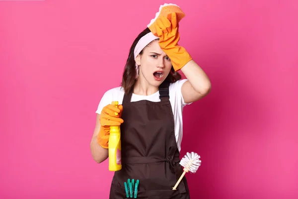 Pembantu rumah tangga wanita yang sedang memegang spons dan deterjen di tangan, mengenakan celemek, karena banyak pekerjaan yang harus dilakukan, terisolasi di atas latar belakang merah muda. Salin spase untuk teks promosi Anda . — Stok Foto