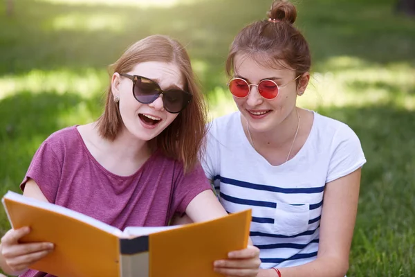Két fiatal szép mosolygó nő olvasó dolog érdekes, ül a parkban, a nyári zöld fű felkészülni vizsga toogether, alkalmi pólók és napszemüveget visel. Oktatás, koncepció diákok. — Stock Fotó