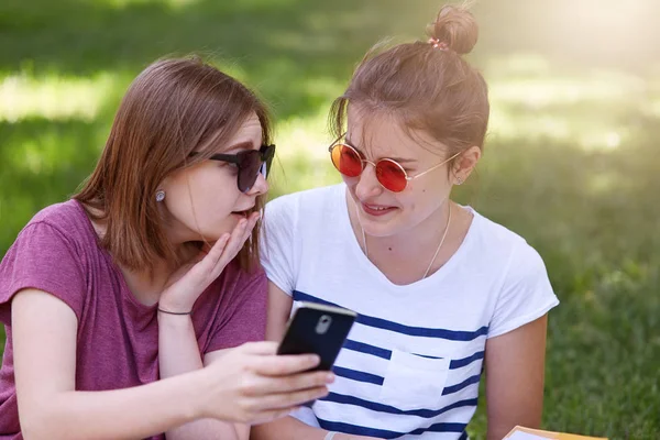 Kép a két lány keresi a szúró telefon, és beszél a park. Vonzó nők pillantást gondolkodó ül a fűben, beszél, új képek a szociális netwoks. Barátság és életmód fogalmak. — Stock Fotó