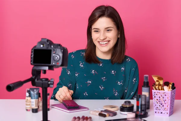 Charming female blogger sits at table with lots cosmetics products, records tutorial video ,looks at her digital camera. Smiling girl talking with her followers, online transliation. Beauty concept.