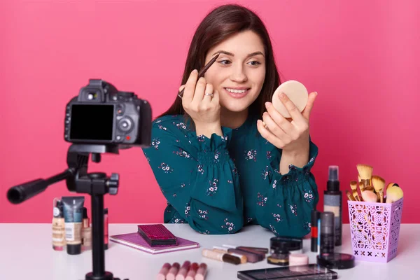 Imagen de la joven bloguera, lleva una blusa elegante, hermosa mujer se sienta frente a la cámara digital, enseña a sus seguidores a aplicar sombra de ojos, transliación en línea, modelo posando aislado sobre fondo rosa — Foto de Stock