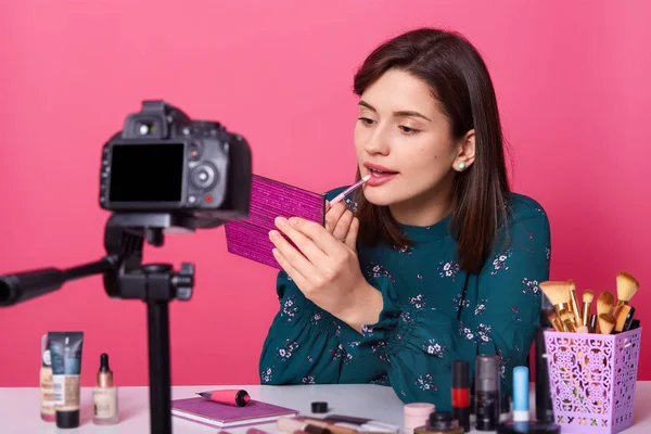 Imagen de una atractiva bloguera haciendo video para el blog de belleza, sentada en una mesa blanca sobre un fondo rosado y aplicando lápiz labial en sus labios mientras mira un pequeño espejo. Concepto de belleza . — Foto de Stock