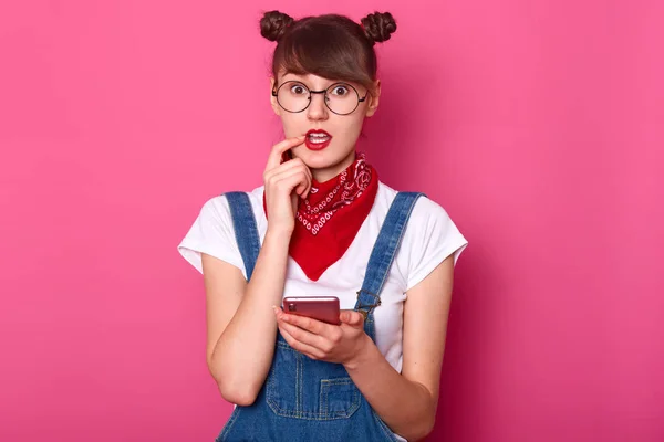 Retrato de chica linda confundida lleva overoles de mezclilla, camiseta blanca casual, bandana roja en el cuello, sostiene el teléfono móvil en la mano, mantiene el dedo delantero en el labio y mirando a la cámara aislada sobre el fondo rosa — Foto de Stock