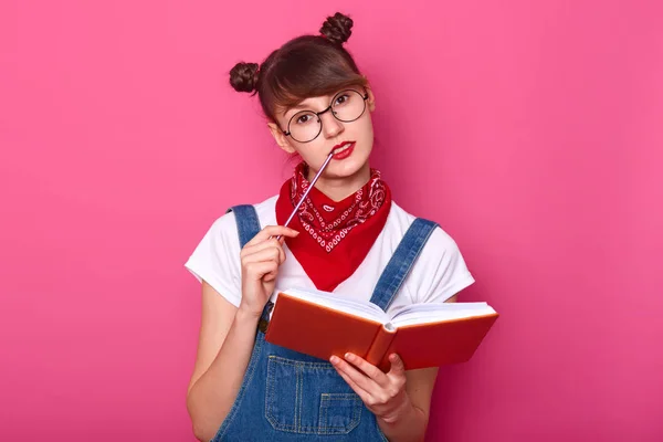 Primer plano retrato de encantador estudiante reflexivo en gafas piensa en algo importante, tiene planificador en las manos, se prepara para los exámenes, mantiene pensil en el labio, posa sobre el fondo rosa . —  Fotos de Stock