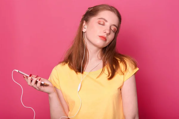 Dreamy perempuan dengan ekspresi termenung dan mata bibit, memiliki headphone modern, mendengarkan musik, menghabiskan waktu luang sendirian, berpose terhadap latar belakang merah muda dengan salinan kosong ruang untuk rata-rata Anda . — Stok Foto